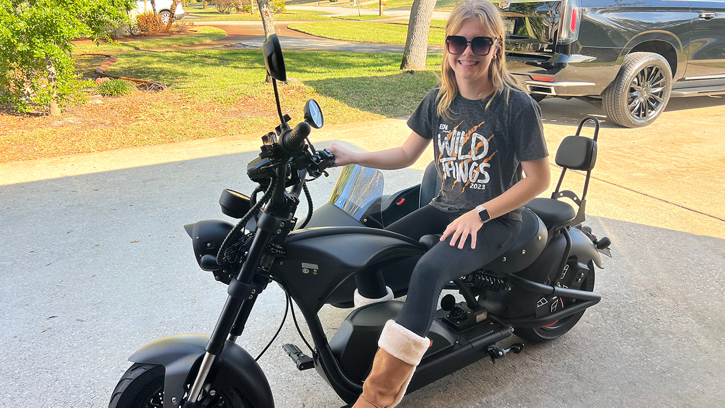 A smiling woman sits on a black chopper-style electric motorcycle with a sidecar